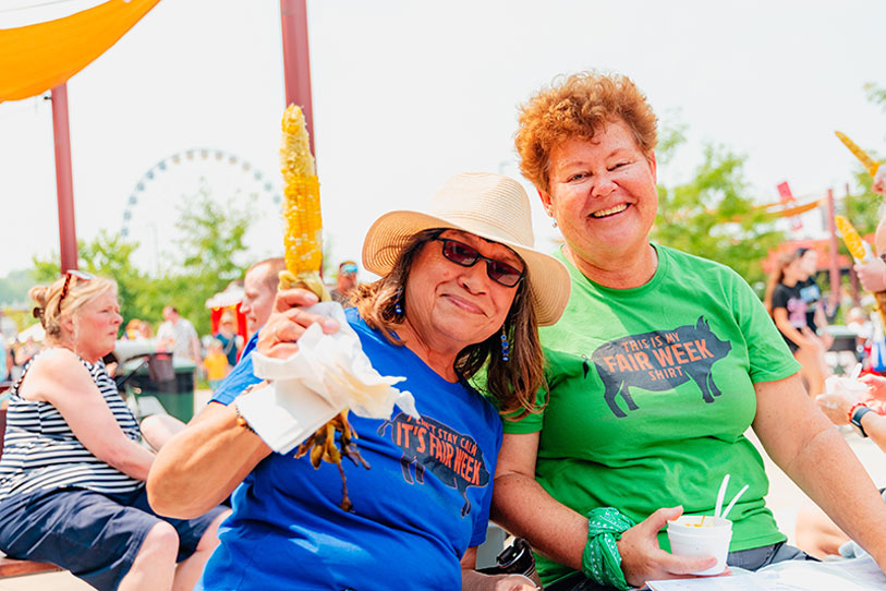 Two women smiling at an outdoor fair, one holding corn on the cob, both wearing “Fair Week” shirts. People and a Ferris wheel are visible in the sunny background.