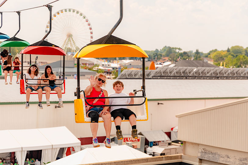 People ride colorful open chairlifts above a fairground. A woman and a child in the front car wave and smile at the camera. In the background, a Ferris wheel and more fair structures are visible under a sunny sky.