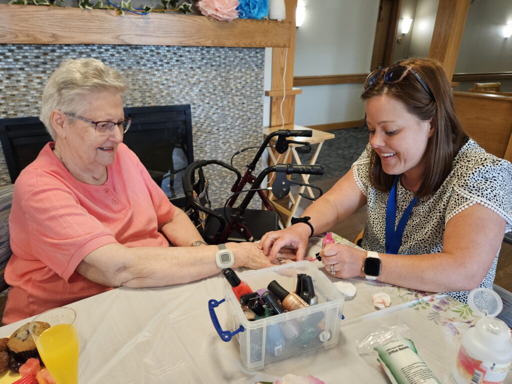 An older woman smiles as a younger woman paints her nails at a table covered with nail polish bottles. They are seated indoors near a fireplace, with a walker visible in the background.