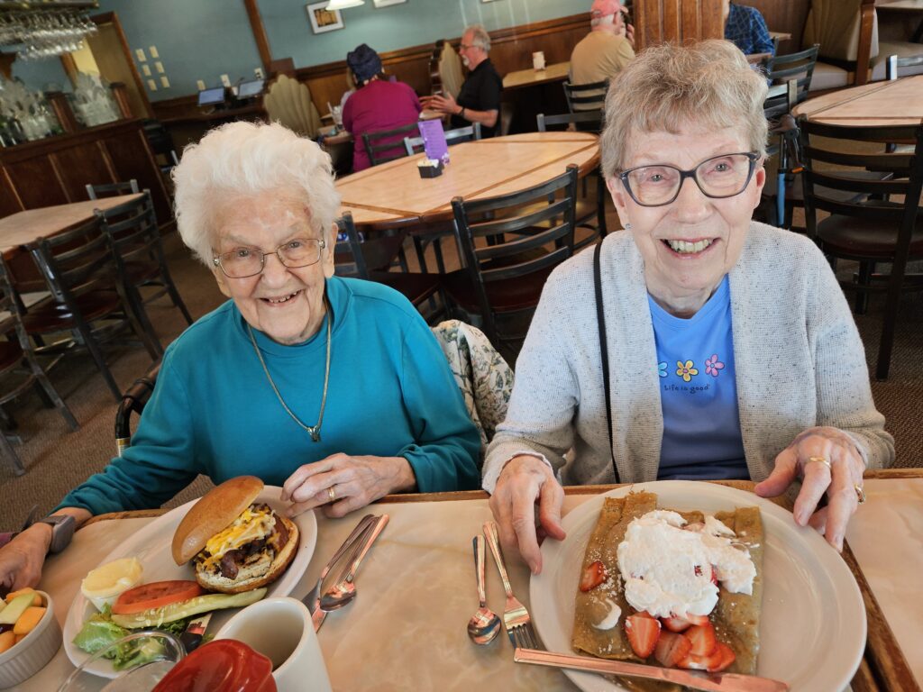 Two elderly women sit together in a restaurant, smiling at the camera. One has a burger with fries, and the other has a crepe topped with whipped cream and strawberries. Both appear happy and are wearing glasses.