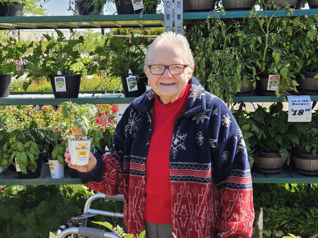 An older woman wearing glasses and a patterned sweater stands outdoors in front of shelves with potted plants, smiling and holding a small plant in one hand. A walker is visible in front of her.