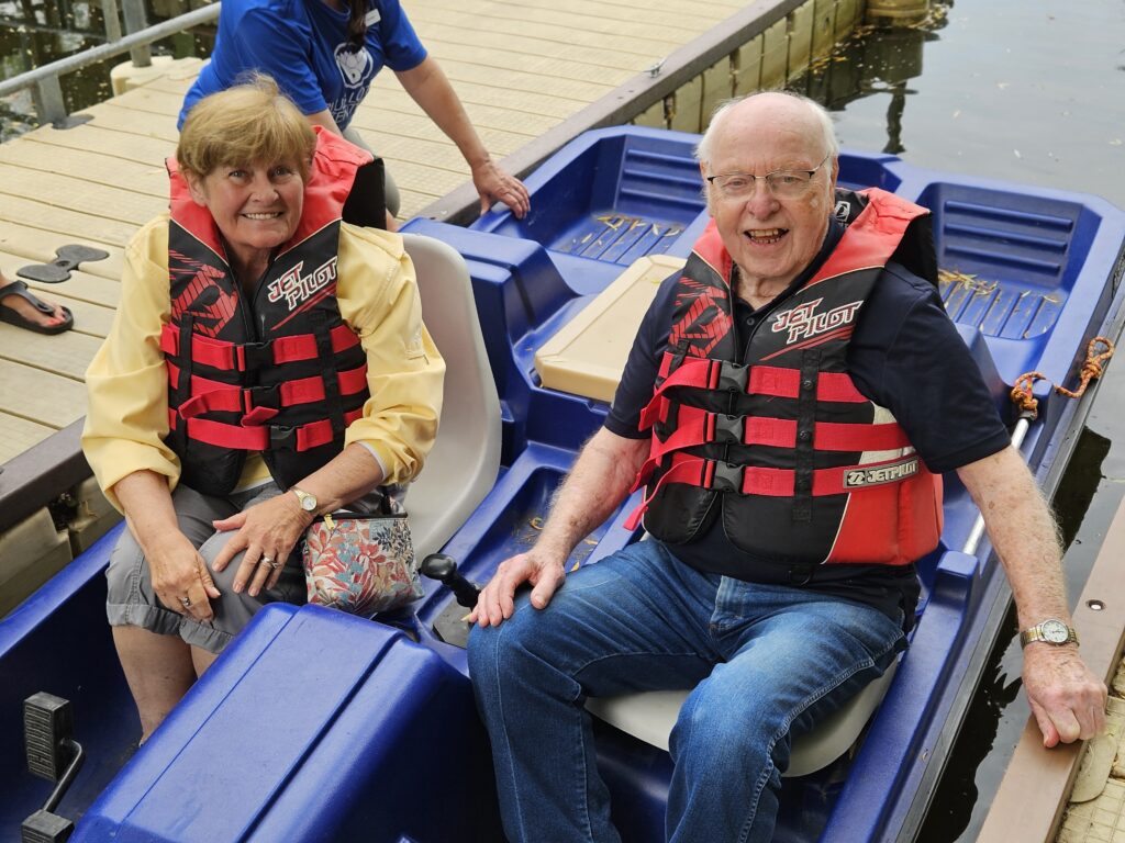 An older woman and man, both smiling and wearing red life vests, sit together in a blue pedal boat at a dock. The woman holds her purse on her lap, and a third person stands behind them on the dock.