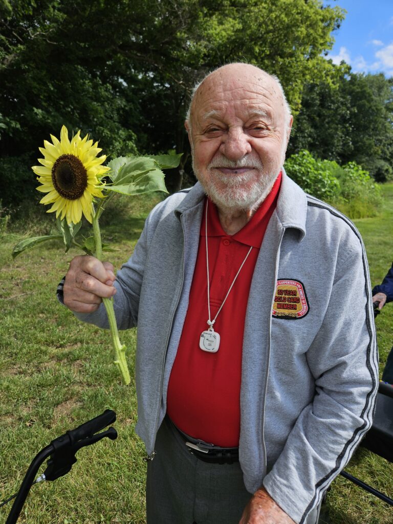 An elderly man with a white beard, wearing a gray jacket and red shirt, smiles while holding a sunflower outdoors on a grassy area with trees in the background.