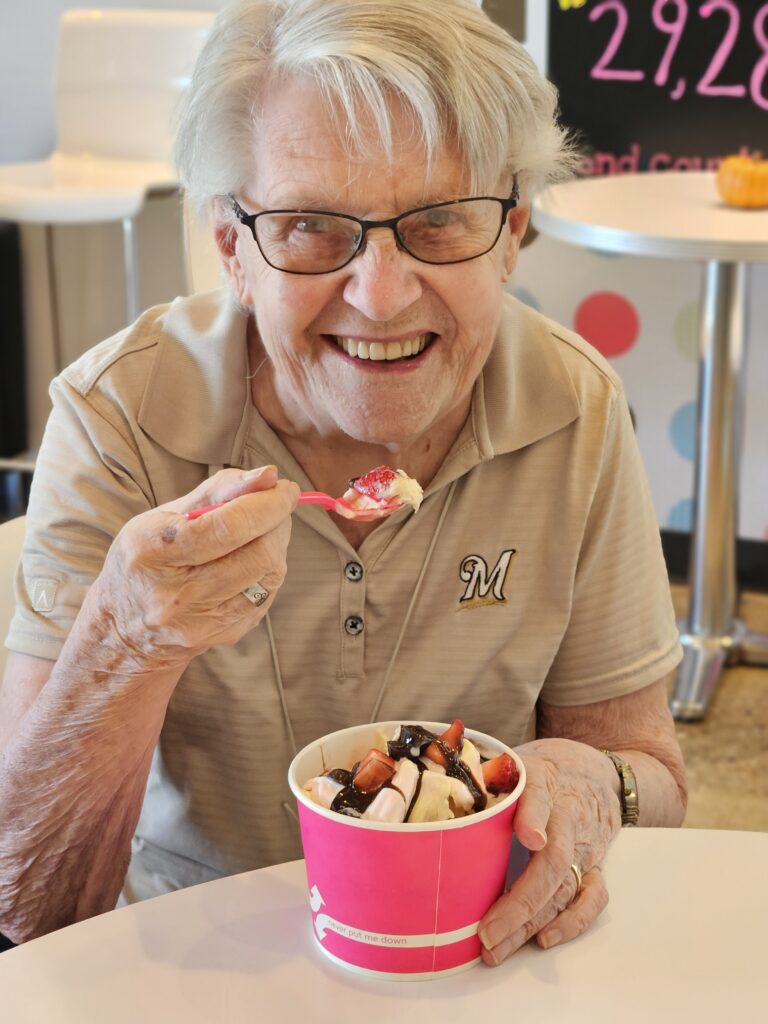 An older woman with glasses smiles while holding a spoon and a large pink cup of ice cream topped with fruit and chocolate syrup. She is seated indoors, wearing a light polo shirt with an M logo.