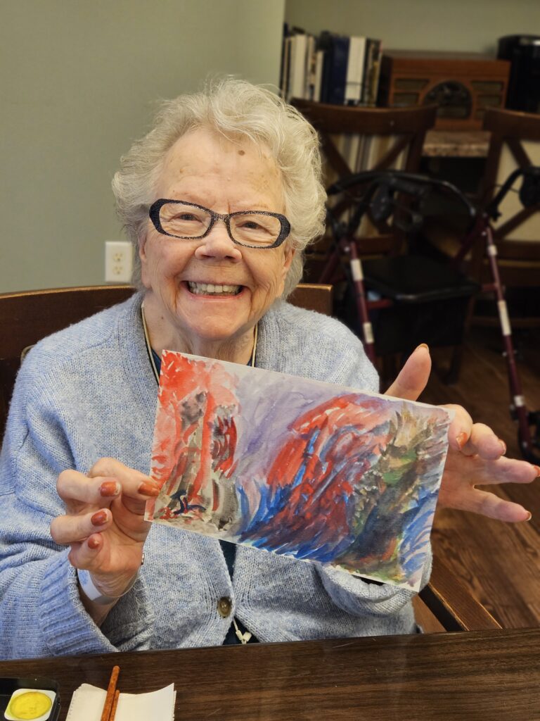 An elderly woman with glasses smiles while holding up a colorful abstract painting she made. She is seated at a table with art supplies, and a walker is visible in the background.