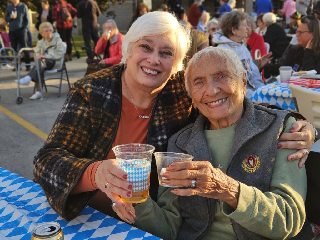 Two older women smiling and holding up drinks sit close together at an outdoor event, surrounded by people at tables with blue-and-white checkered tablecloths, enjoying a festive, sunny atmosphere.