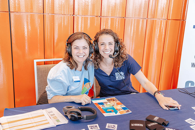 Two women wearing headsets sit at a blue table with papers and promotional items, smiling at the camera. An orange wall is behind them, and they appear to be hosting or recording an event or podcast.