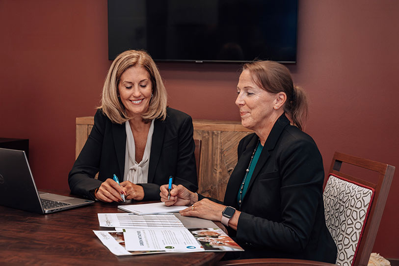 Two women in business attire sit at a table with documents and a laptop, smiling and writing notes. They appear to be having a professional meeting in an office with a maroon wall and a TV in the background.