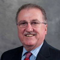 A middle-aged man with short gray hair, glasses, and a mustache smiles at the camera. He is wearing a dark pinstripe suit, a light blue shirt, and a red patterned tie. The background is a plain gray.