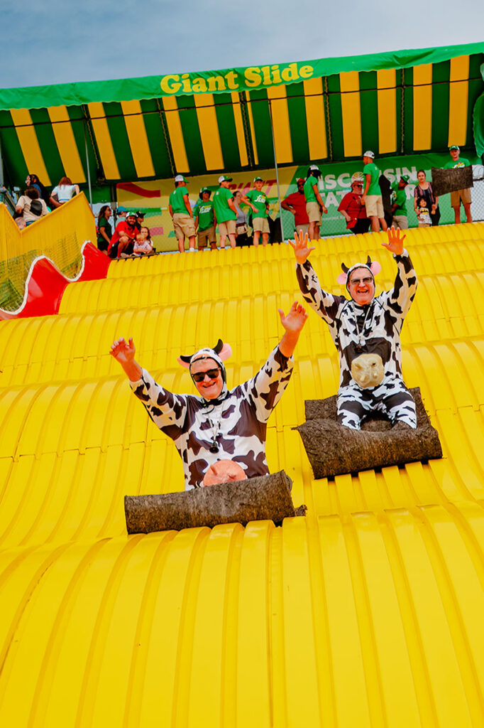 Two people dressed in cow costumes slide down a large, bright yellow slide on mats, with their arms raised. In the background, a crowd of people watches from behind a fence under a green-and-yellow canopy labeled Giant Slide.