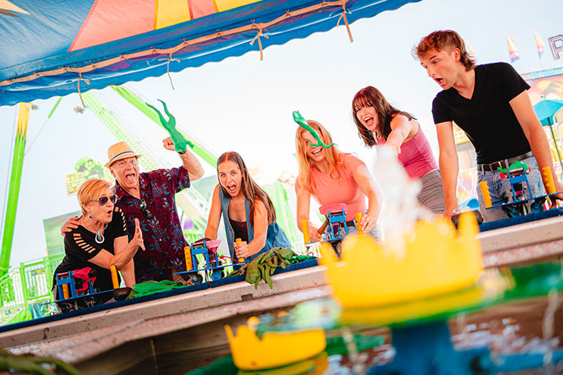 Six excited people cheer and compete in a water gun race game at a colorful amusement park booth, with bright rides visible in the background and yellow crowns as game targets in the foreground.