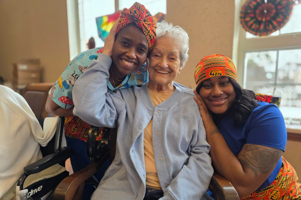 Three women smile and pose together indoors. The women on each side wear colorful headwraps and lean their heads against the elderly woman in the center, who is wearing a light blue cardigan. Sunlight streams through windows behind them.