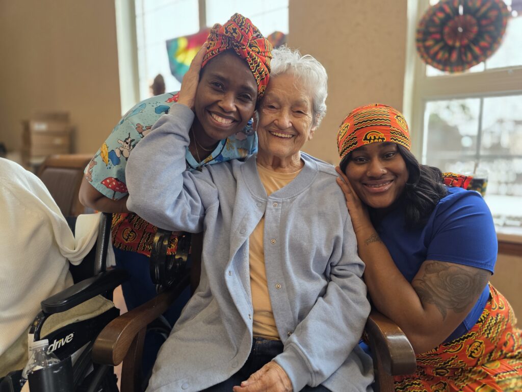Three women sit closely together, smiling warmly. The woman in the center is elderly and wears a light blue cardigan. The two women on either side wear colorful headwraps and gently hug her, creating a joyful and loving atmosphere.