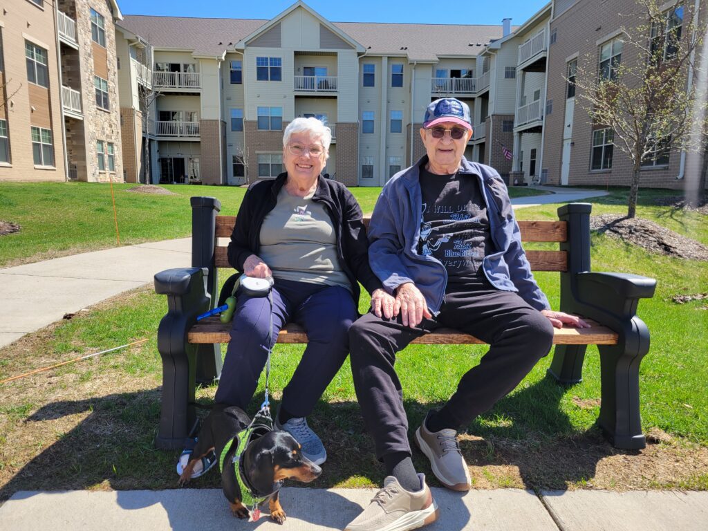 An elderly woman and man sit together on a wooden bench outside an apartment building on a sunny day, smiling. A small black and brown dog on a leash sits at their feet on the grass.