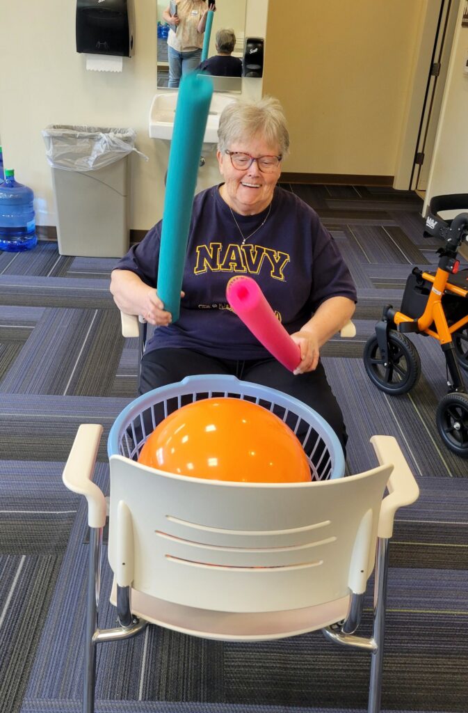 An older woman wearing a Navy t-shirt smiles while sitting in a chair, holding two colorful foam sticks and playing an orange ball in a basket placed on another chair. A walker and water cooler are visible nearby.