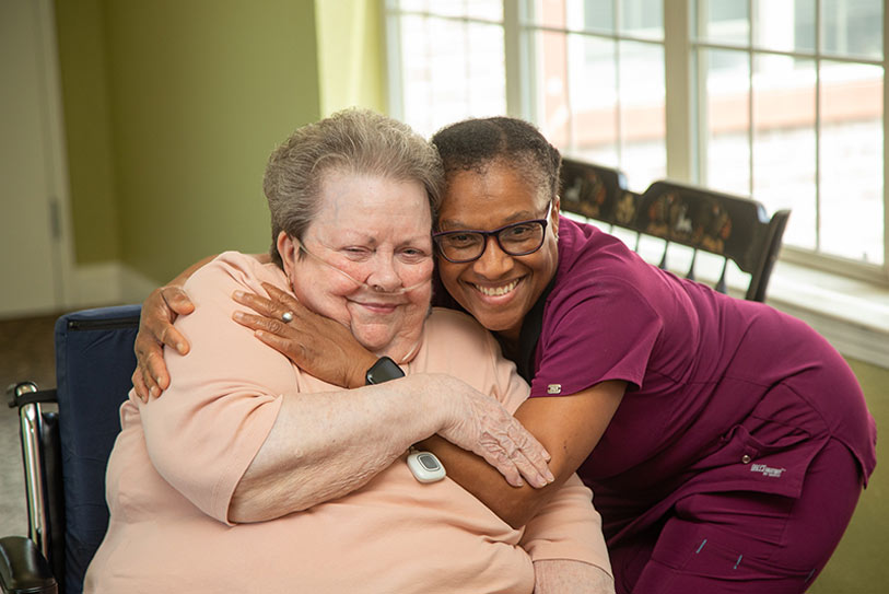 An elderly woman in a wheelchair smiles as she is hugged by a caregiver in purple scrubs. They are indoors by a window, both looking happy and content.