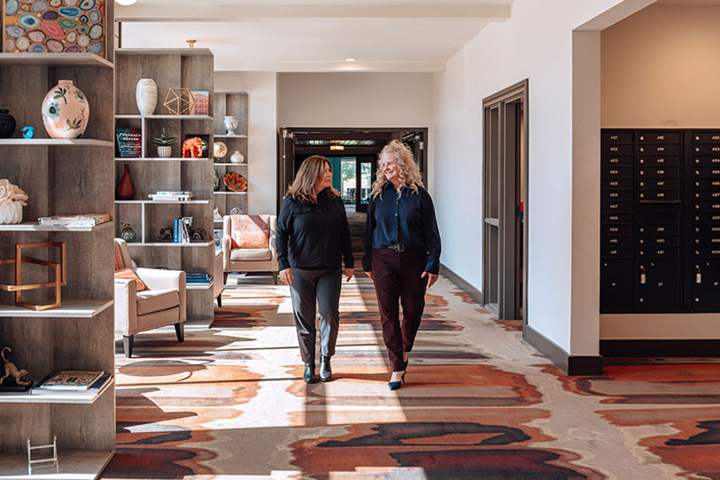 Two women walk side by side in a modern, brightly lit hallway with stylish decor, bookshelves, armchairs, and mailboxes lining the walls. The space has a patterned carpet and contemporary furnishings.
