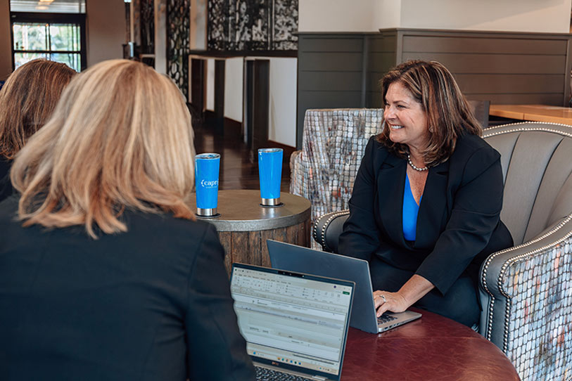 Two women sit at a round table with laptops open, engaged in conversation. One woman smiles while seated on a couch; two blue travel mugs are on the table. The setting appears to be a modern, open office space.
