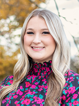 A young woman with long, blonde hair smiles outdoors. She is wearing a black top with pink and green floral patterns. Fall foliage and a cloudy sky are visible in the blurred background.