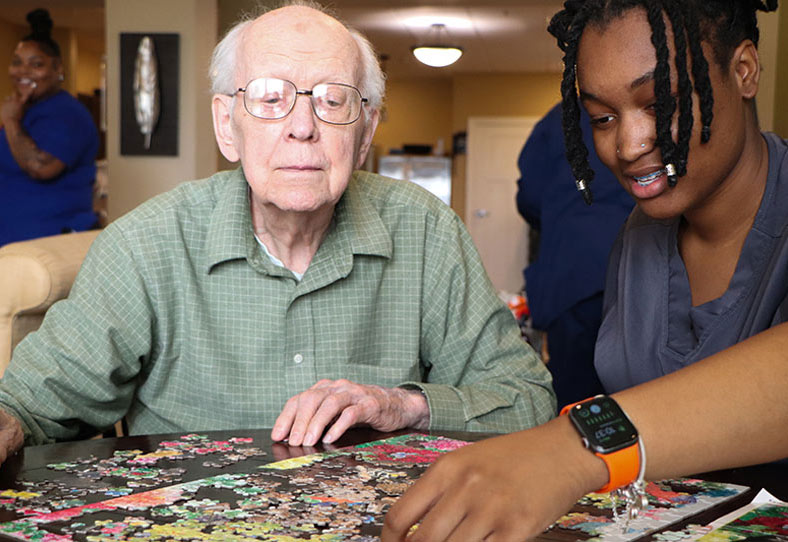 An elderly man and a young woman work together on a jigsaw puzzle at a table. The man wears glasses and a green shirt; the woman, in dark clothing and wearing a smartwatch, reaches to place a puzzle piece. A smiling person is in the background.