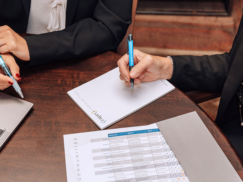 Two people in business attire sit at a table with one writing on a notepad and the other holding a pen. A printed chart with checkboxes and a folder are also visible on the table.