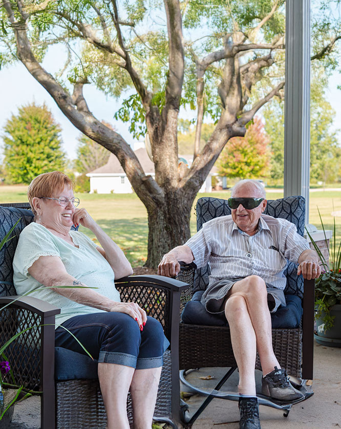 An elderly woman and man sit outside on a patio, smiling and talking. The man is wearing dark sunglasses and both are seated in cushioned chairs with trees and a lawn in the background.