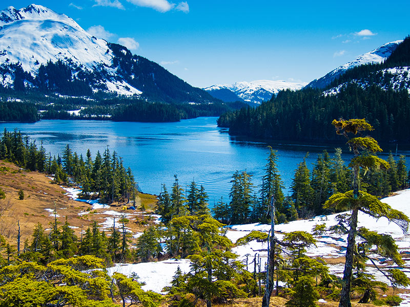 Snow-capped mountains surround a clear blue lake bordered by dense evergreen forests and patches of snow, under a bright blue sky with scattered clouds.