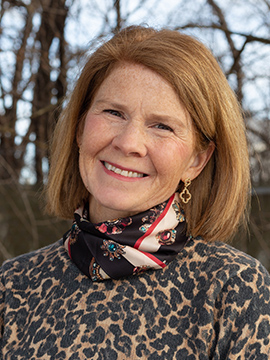 A middle-aged woman with straight, light brown hair smiles outdoors. She is wearing a leopard-print top and a patterned scarf, with bare trees visible in the background.