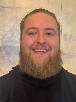 A smiling person with a full beard and long hair pulled back, wearing a textured dark sweater, stands in front of a beige and brown marbled background.