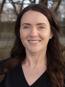 A woman with long brown hair and blue eyes is smiling at the camera outdoors. She is wearing a black top, and blurred trees are visible in the background.