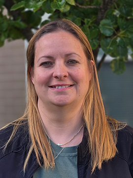 A woman with straight, shoulder-length blonde hair smiles at the camera. She is wearing a dark jacket over a green top and a necklace, standing outdoors with green leaves and a blurred building in the background.