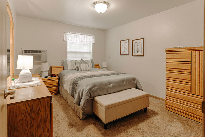 A cozy bedroom with a double bed featuring gray and white bedding, a wooden dresser and chest of drawers, a cushioned bench at the bed’s foot, beige carpet, and framed art on the white walls.