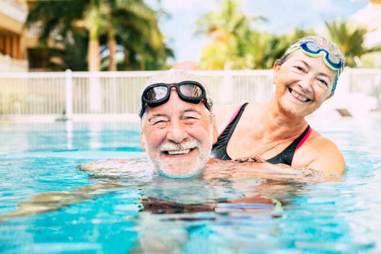 An older man and woman wearing swim goggles smile and enjoy themselves in an outdoor swimming pool, with palm trees and buildings visible in the background.
