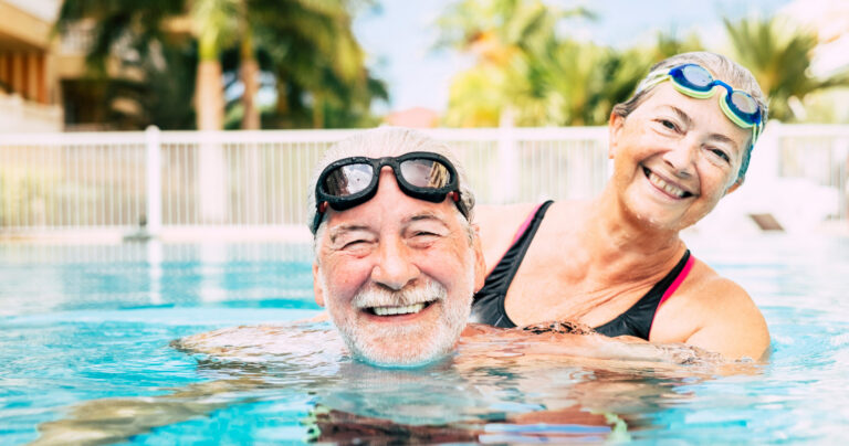 An older man and woman wearing swim goggles smile and enjoy themselves in an outdoor swimming pool, with palm trees and buildings visible in the background.