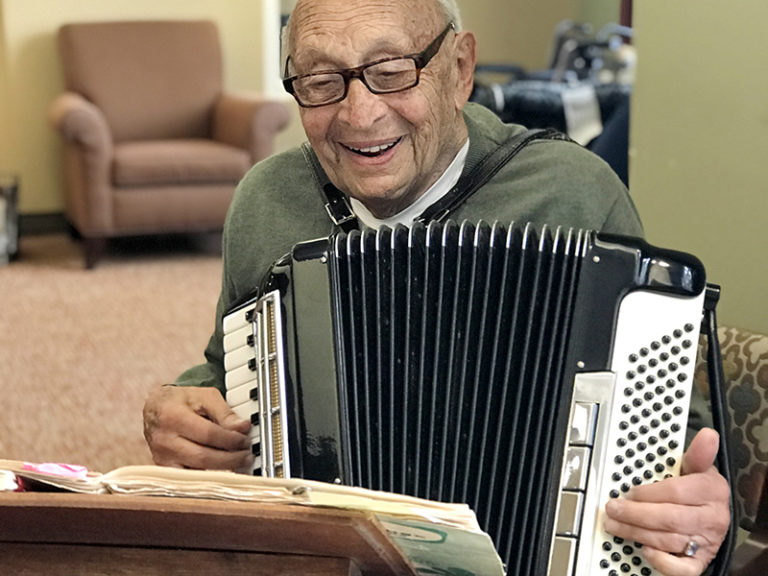 An elderly man wearing glasses and a green sweater smiles while playing an accordion. He sits at a table with sheet music, with a chair and soft lighting in the background.