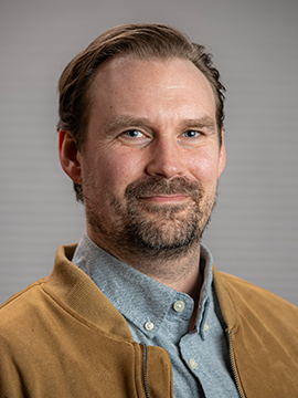 A man with light skin, short brown hair, and a beard is smiling slightly. He is wearing a tan jacket over a gray collared shirt. The background is plain and softly blurred.