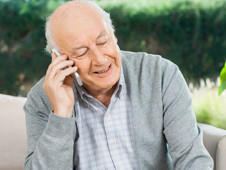 An elderly man with a bald head and gray sweater smiles as he talks on a smartphone, sitting on a light-colored sofa with greenery visible in the background.