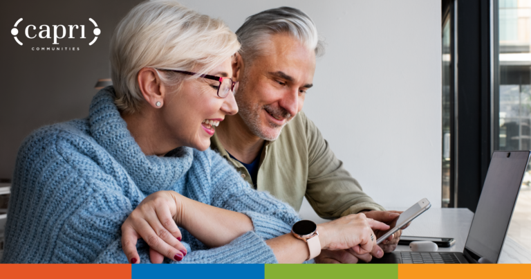 An older woman and man smile while looking at a smartphone together in front of a laptop. The woman wears glasses and a blue sweater. A Capri Communities logo is in the top left corner.