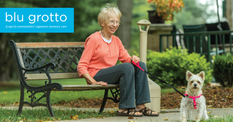 An elderly woman with glasses sits smiling on a park bench, holding a small dog on a leash. The dog stands on the path beside her. Greenery and a house are in the background. blu grotto text appears in the top left corner.