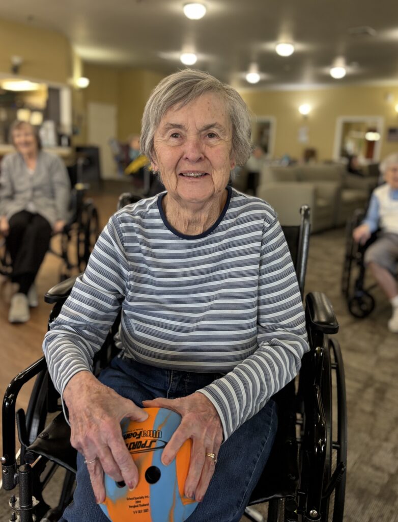 An elderly woman in a wheelchair smiles while holding a small orange and blue ball. She wears a striped shirt, and other seniors are visible in the background in a bright, communal room.