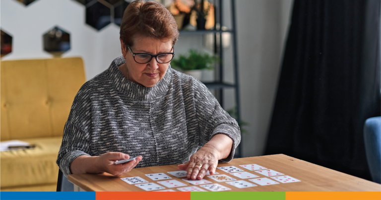 An older woman with short hair and glasses sits at a table, laying out playing cards in a grid pattern. She is focused on the cards, and the setting is a cozy room with modern decor.