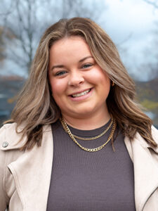 A woman with wavy light brown hair smiles at the camera. She is wearing a beige jacket over a dark top and layered gold necklaces. The background is outdoors with blurred trees and a cloudy sky.
