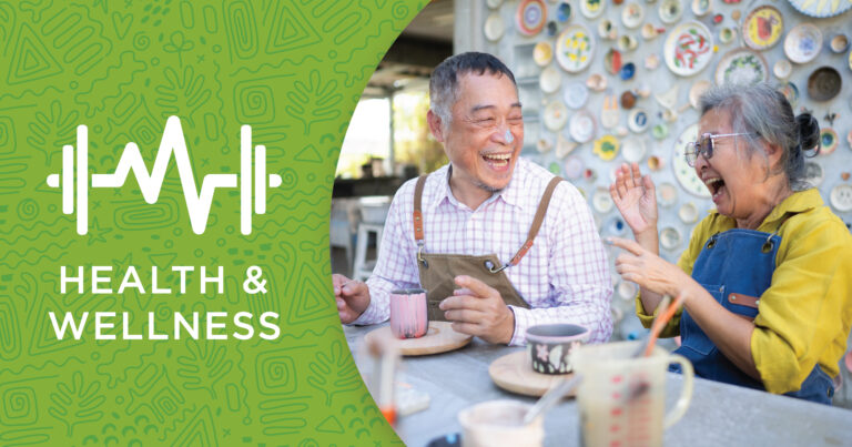 Two older adults laugh together at a table with mugs, set against a wall decorated with plates. On the left, a green graphic reads “Health & Wellness” with a heartbeat and dumbbell icon.