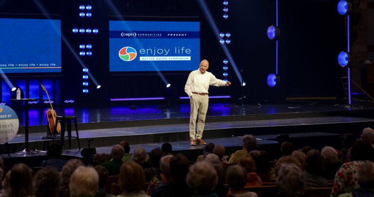 A man stands on stage speaking to an audience at the “Enjoy Life: Active Aging Symposium,” with bright lights and a large screen displaying the event name behind him.