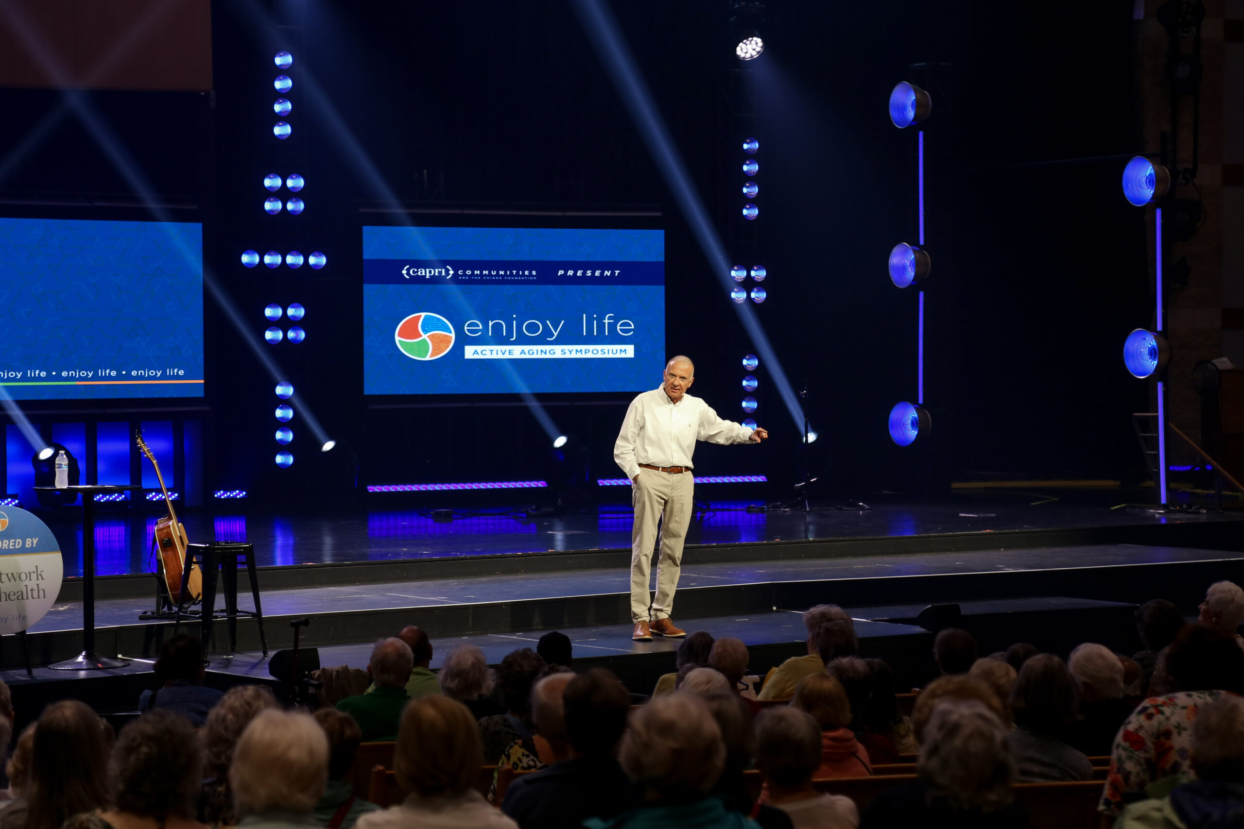 A man stands on stage speaking to an audience at the “Enjoy Life: Active Aging Symposium,” with bright lights and a large screen displaying the event name behind him.