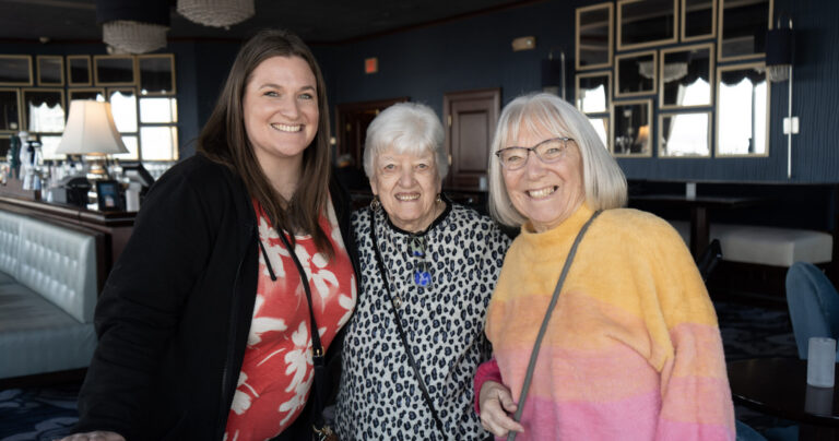 Three women stand together indoors, smiling at the camera. One wears a red and white top, another a leopard print top, and the third a yellow and pink sweater. The background shows tables, chairs, and large mirrors.