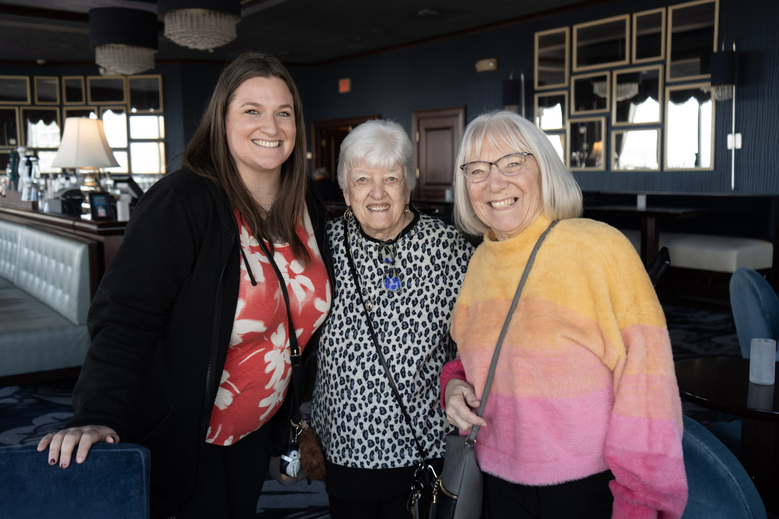 Three women stand together indoors, smiling at the camera. One wears a red and white top, another a leopard print top, and the third a yellow and pink sweater. The background shows tables, chairs, and large mirrors.