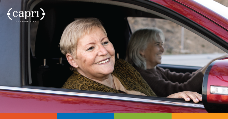 Two older women smiling while sitting inside a red car, with one in the driver’s seat and the other in the passenger seat. The Capri Communities logo is in the top left corner. A colorful bar is at the bottom of the image.