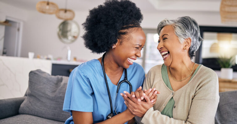 A nurse in blue scrubs and an elderly woman sit on a couch, holding hands and laughing joyfully together in a bright living room. The mood is warm, friendly, and caring.