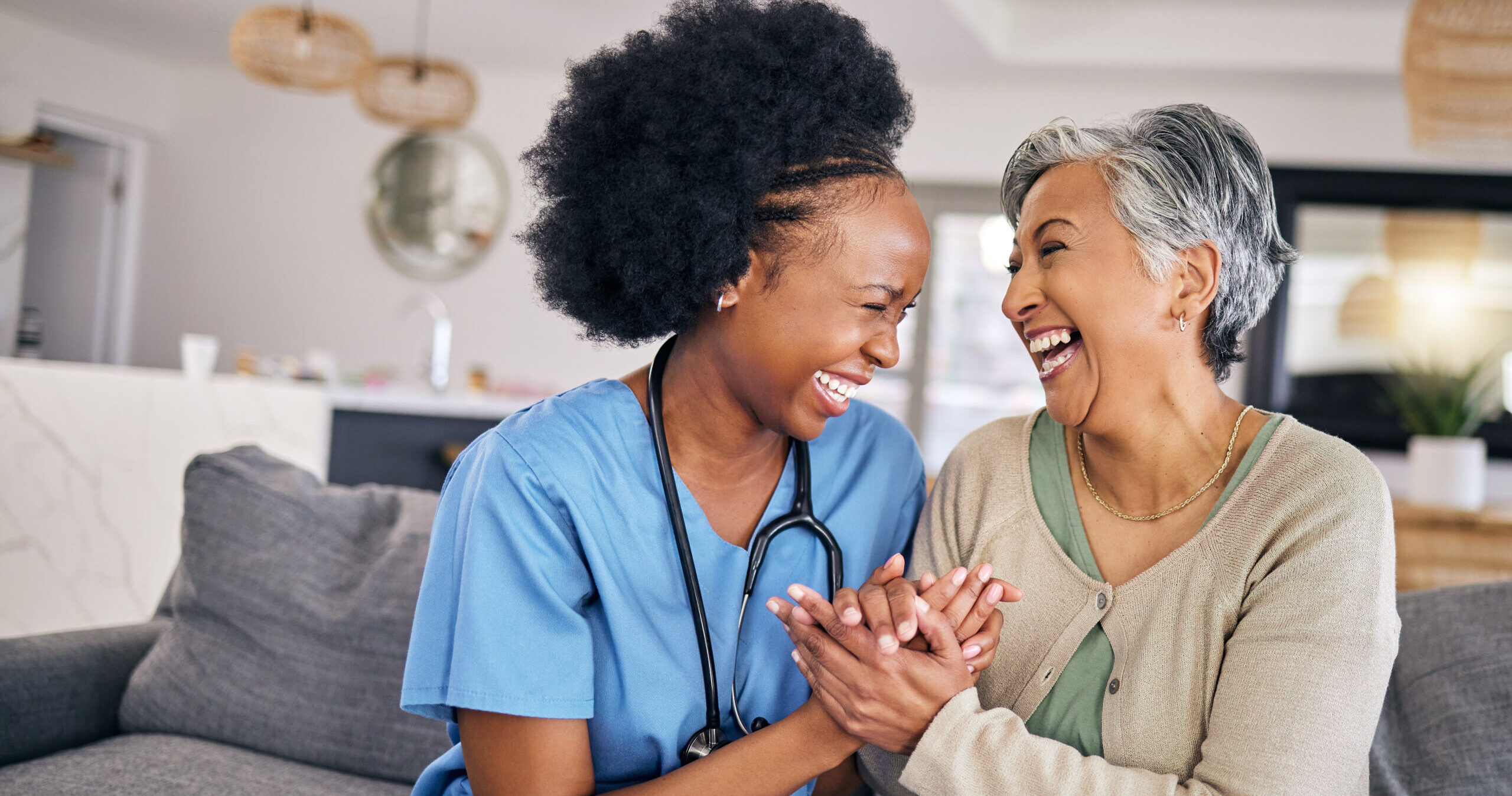 A nurse in blue scrubs and an elderly woman sit on a couch, holding hands and laughing joyfully together in a bright living room. The mood is warm, friendly, and caring.